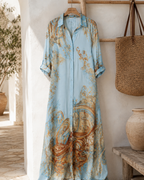 The dress on a wooden hanger against a white wall, showing the full length, the button front from collar to hem and the tabs that hold the rolled sleeves.