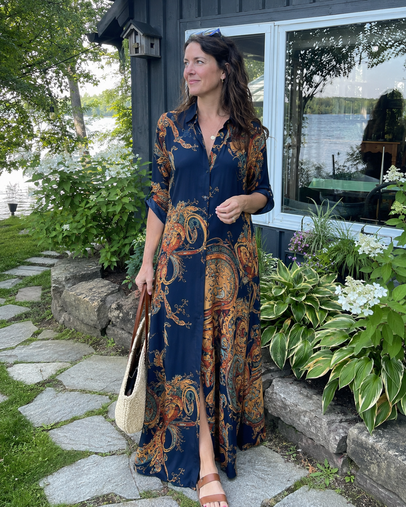 Customer photo: the navy dress worn on a stone path beside a lake house, sleeves rolled, carrying a straw bag.