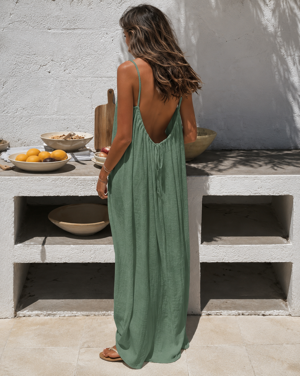 Woman wearing the Cleo strappy maxi dress in sage green, standing in a whitewashed courtyard