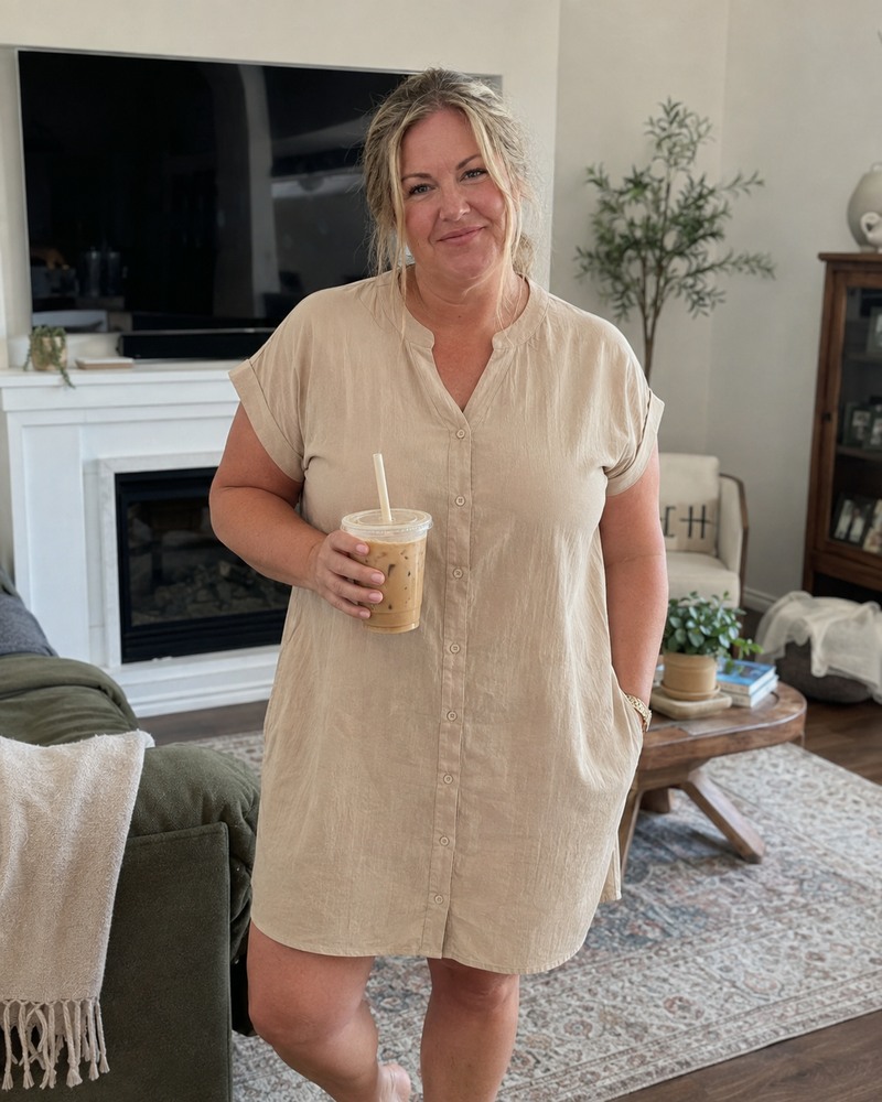 Woman wearing the Odette shirt dress in beige, standing in a living room with an iced coffee
